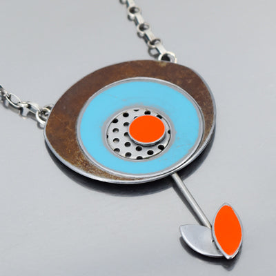 Maeve - Ford & Oldsmobile: A necklace with a circular pendant of bronze, turquoise, and oxidized sterling silver (red dot center), plus a thin stem ending in an orange and silver leaf.