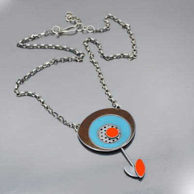 The Maeve - Ford & Oldsmobile necklace features a silver chain and a modern pendant with concentric circles in brown, blue, oxidized sterling silver, and an orange center dot, plus a silver stem with orange and silver leaf below.
