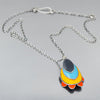 The Rhoda - Ford is a sterling silver chain necklace with a pendant of layered oval shapes in dark gray, blue, yellow, and orange, forming a stylized flower or abstract design, shown on a gray surface. Made with vintage car parts by Joyride Jewelry
