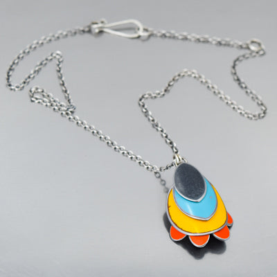 The Rhoda - Ford is a sterling silver chain necklace with a pendant of layered oval shapes in dark gray, blue, yellow, and orange, forming a stylized flower or abstract design, shown on a gray surface. Made with vintage car parts by Joyride Jewelry