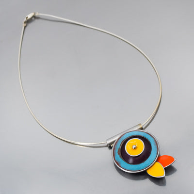 The Verna - Ford & Mercury is a silver necklace on a sterling cable featuring a stylized fish pendant with concentric blue, yellow, and purple circles, plus orange, yellow, and white teardrop fins. Pendant dimensions available upon request.
