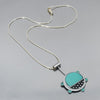 The Gertrude - Chevy & Ford is a  necklace with a round turquoise pendant, black dotted accents, and small blue tabs around the edge, hanging from a sterling silver chain on a light gray surface. Handmade one of a kind design by Joyride Jewelry.