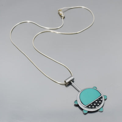 The Gertrude - Chevy & Ford is a  necklace with a round turquoise pendant, black dotted accents, and small blue tabs around the edge, hanging from a sterling silver chain on a light gray surface. Handmade one of a kind design by Joyride Jewelry.