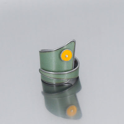 The Marlin - Chevy & Ford is a modern green ring with an asymmetrical design, featuring a yellow circular accent made with  original car parts by Joyride Jewelry, shown on a reflective gray surface.