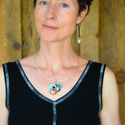 A woman with short brown hair models the Alice - Chevy & Pontiac set: a black sleeveless top, sterling silver ear wires with turquoise and orange earrings, and a matching circular pendant necklace, all against a wooden background.