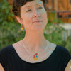 A woman with short brown hair in a black top poses outdoors, wearing the whimsical Wilma - Ford & Pontiac red pendant necklace and matching bold, colorful earrings. In the background, a blurred Pontiac Tempest sits among lush greenery.