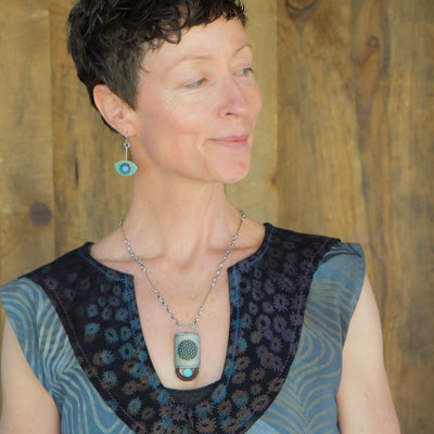 A woman with short dark hair wears the Eula - Dodge, Pontiac, Chevy: a sleeveless patterned blue top paired with a silver necklace and matching earring. She stands before a wooden background, gazing to the side with a soft smile.