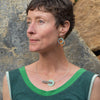 A woman with short brown hair wears a sleeveless green top and modern geometric jewelry, including Evelyn - Ford & Chevy earrings with sterling silver ear wires. She stands outdoors by large textured rocks, glancing to the side.
