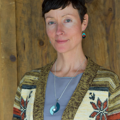 A woman with short brown hair, wearing the Shirley - Ford & Dodge turquoise earrings and matching necklace on a sterling silver chain, stands in front of a wooden wall, looking at the camera with a slight smile.