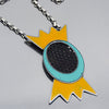 The Wonda - Datsun & Chevy is a pendant necklace with a geometric design, black oval center with small holes, turquoise border, bright yellow angular shapes, on an oxidized sterling silver chain.