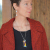 A woman with short hair models the Gracie - Ford & Dodge textured red coat, black top, round silver earrings, and a colorful geometric pendant on an oxidized sterling silver chain against a wooden background.