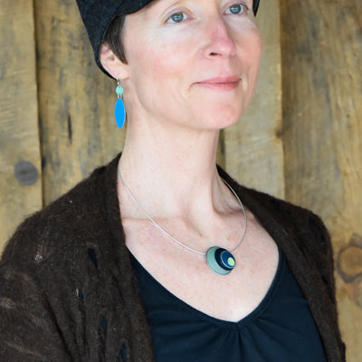 A woman wearing the Lenora - Ford & Dodge round black and blue pendant necklace with a sterling silver cable stands in front of a wooden background, also dressed in a black hat, blue earrings, black shirt, and brown sweater.