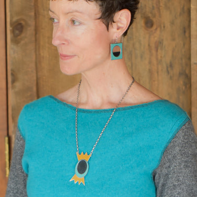A woman with short hair wears the Wonda - Datsun & Chevy: a turquoise top and matching geometric jewelry—pendant necklace on oxidized sterling silver and square earrings in teal, black, and yellow—standing by a wooden wall.