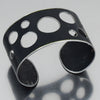 Elwood - GMC pickup: A sleek, wide black metal cuff bracelet featuring various-sized circular cutouts, showcased on a reflective surface.