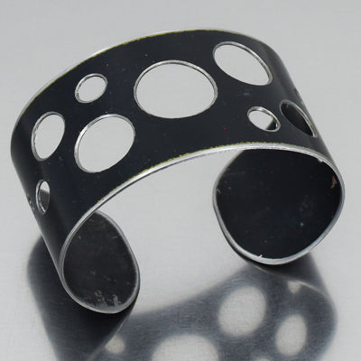 Elwood - GMC pickup: A sleek, wide black metal cuff bracelet featuring various-sized circular cutouts, showcased on a reflective surface.
