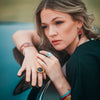 A woman with wavy blonde hair leans on a glossy Della - AMC & Ford car, gazing thoughtfully into the distance. She wears statement rings, geometric earrings, and a black top that contrasts beautifully with the car.