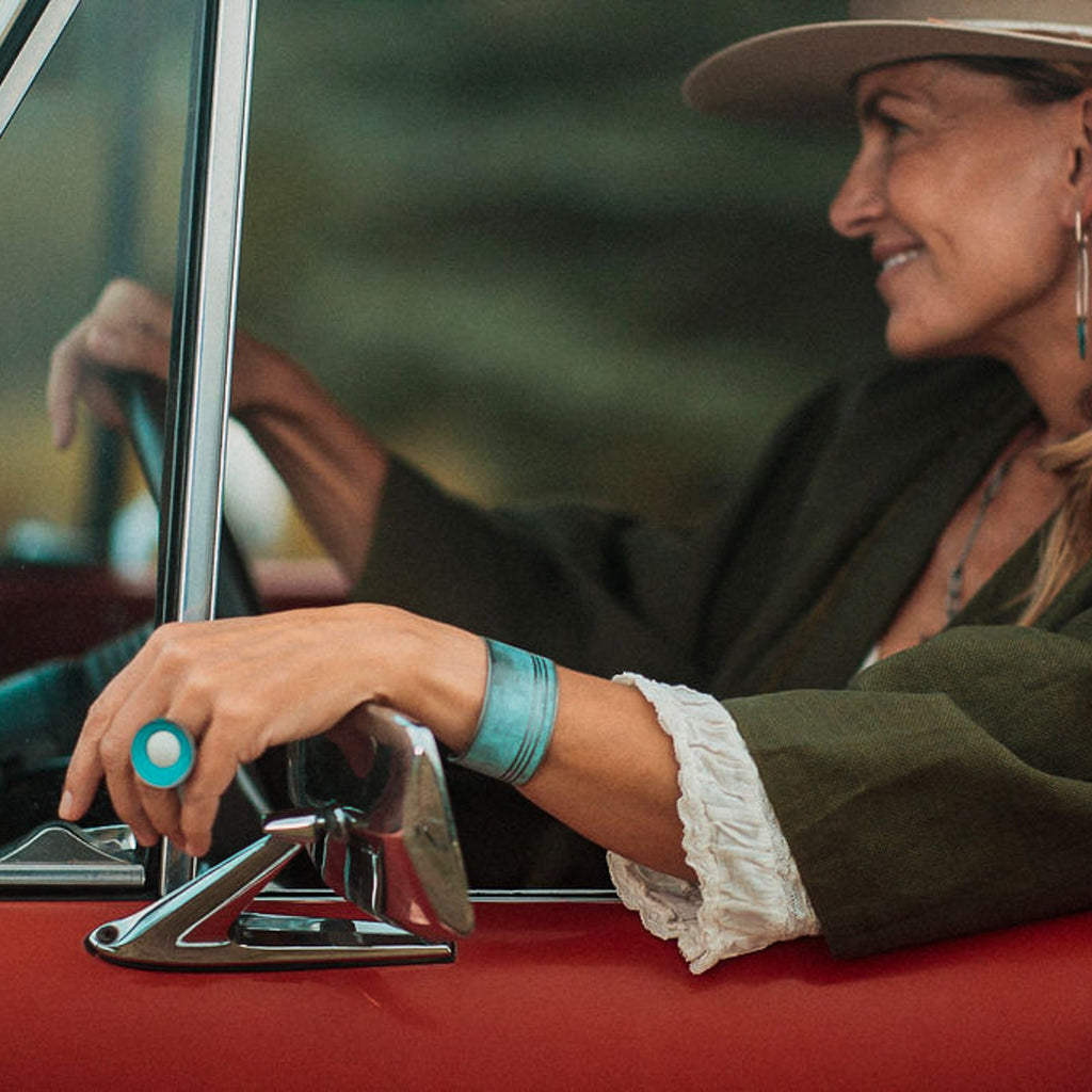 A smiling woman in a hat and green jacket sits in the driver’s seat of  a 74 Ford Bronco wearing the Frankie cuff bracelet and a ring. Each piece is made from vintage cars with the original car paint intact.