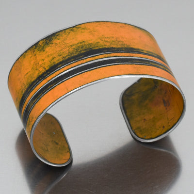 The Frankie - Dodge pickup is a wide metal cuff bracelet with a matte orange enamel finish made with classic car parts, accented by two thin black stripes. This adjustable bracelet is shown on a reflective gray surface.