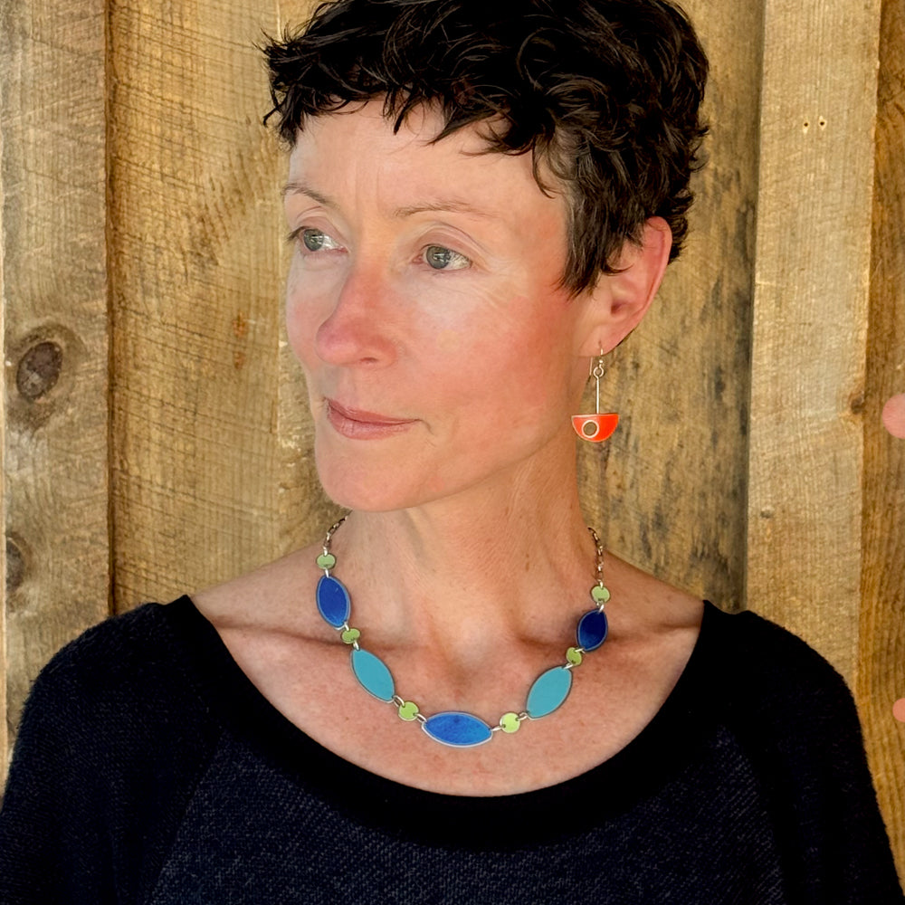 Woman modeling jewelry, wearing a bright, colorful, unique, original necklace and earrings.
handmade using vintage car parts with the original car paint intact. Joyride Jewelry. Jewelry made from vintage car parts. sustainable, recycled 

