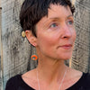 Woman wearing Wilma earrings featuring orange 79 International Scout car paint and sterling silver ear wires against wood background