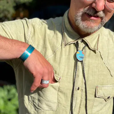 A man in a light green shirt, bolo tie with a colorful round pendant, adjustable ring, and blue bracelet slips his hand inside his chest pocket. His face is partly seen. wearing jewelry by Joyride Jewelry.
