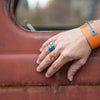A hand wearing the Joey - Chevy & AMC blue and orange rings and an orange bracelet rests on the rusty door of a vintage vehicle with faded paint, while a blurred outdoor scene through the window hints at its bygone stories.