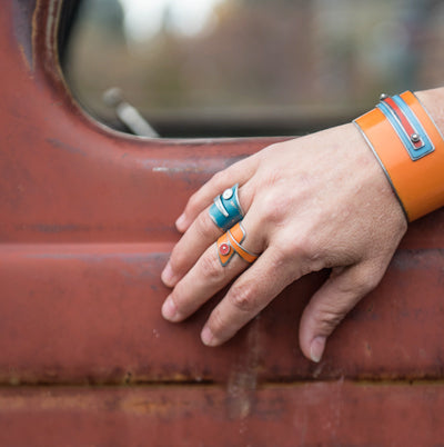 A hand wearing the Joey - Chevy & AMC blue and orange rings and an orange bracelet rests on the rusty door of a vintage vehicle with faded paint, while a blurred outdoor scene through the window hints at its bygone stories.