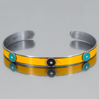 The Emril - Ford & Oldsmobile bracelet showcases  vivid yellow enamel band, two turquoise circular inlays on each side, and a central black circle, displayed on a reflective surface. by Joyride Jewelry