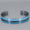 The Jo - Oldsmobile is a metallic cuff bracelet made from vintage car parts with a matte finish in bold turquoise . Its open-ended, adjustable design is showcased on a reflective gray surface. 