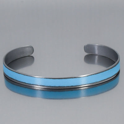 The Jo - Oldsmobile is a metallic cuff bracelet made from vintage car parts with a matte finish in bold turquoise . Its open-ended, adjustable design is showcased on a reflective gray surface. 