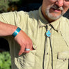 A man wearing a light green button-up shirt, turquoise bracelet, adjustable ring, and bolo tie with a colorful pendant reaches into his pocket . His face is partially visible. All the jewelry is made by Joyride Jewelry.