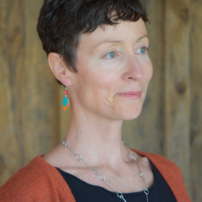 A woman with short brown hair, in a burnt orange cardigan and black top, wears the Willa - Mercury, AMC, Dodge blue and orange earrings made from vintage car parts. She stands before a wooden background, smiling softly.