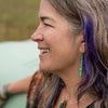 Judith - Ford is shown in profile, smiling outdoors with a natural blurred background. She has long gray hair with a purple streak and wears turquoise jewelry featuring sterling silver ear wires, including matching earrings and necklace.