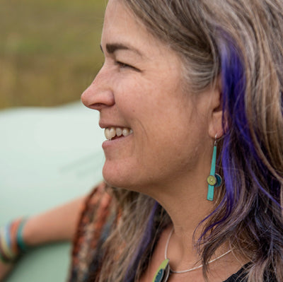 Judith - Ford is shown in profile, smiling outdoors with a natural blurred background. She has long gray hair with a purple streak and wears turquoise jewelry featuring sterling silver ear wires, including matching earrings and necklace.