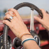 Close-up of two hands wearing rings and an adjustable bracelet, gripping the steering wheel of a vintage red car named Jo - Oldsmobile, with a blurred background visible through the windshield.