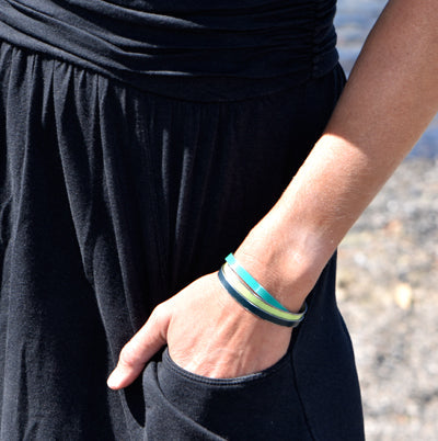 Wearing a black dress with one hand in their pocket, a person showcases the Oliver- Plymouth Satellite adjustable bracelet in green, blue, and black against a blurred outdoor background.