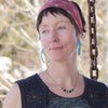 A woman with short brown hair wears a red headband and blue Plymouth Satellite hoop earrings by Claire with stainless steel ear wires. She stands outdoors in a sleeveless top, gazing aside, blurred trees and a chain behind her.
