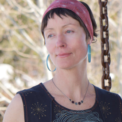 A woman with short brown hair wears a red headband and blue Plymouth Satellite hoop earrings by Claire with stainless steel ear wires. She stands outdoors in a sleeveless top, gazing aside, blurred trees and a chain behind her.