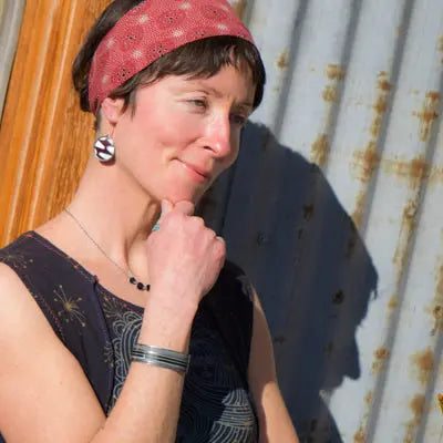 A woman in a red headscarf, patterned earrings, and sleeveless top stands thoughtfully by a corrugated metal wall, sunlight casting her shadow—and the Wilson - Jeep Willys—on the wall.