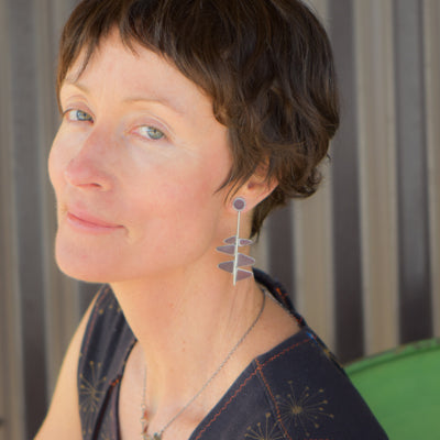 A woman with short brown hair and light skin smiles subtly in front of a green chair and striped background, wearing a sleeveless patterned top, sterling silver geometric earrings, and posing beside Thelma - GMC pickup.