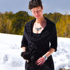 A woman in a black dress stands on snowy ground, smiling with eyes closed as she gently pets a  black dog. Behind her, trees and a cloudy sky. She is modelling a black and white necklace by Joyride Jewelry