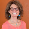 A smiling woman with shoulder-length brown hair, round glasses, and silver necklaces wears Isabel - Ford turquoise and blue earrings with sterling silver ear wires, a pink top, and stands before an orange-brown wall.