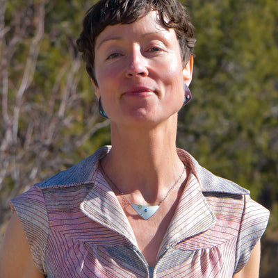 A woman with short brown hair stands outdoors, smiling softly. She wears the Lizzie - Ford necklace—a geometric design on an oxidized sterling silver chain—paired with a sleeveless, collared, pink-striped shirt. Sunlit greenery fills the blurred background.