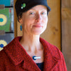 Wearing the Lana - Ford & Dodge necklace, a woman in a textured red coat, black hat with green pins, and blue geometric earrings stands indoors, smiling softly with vibrant artwork blurred behind her. Handmade jewelry by Joyride Jewelry.
