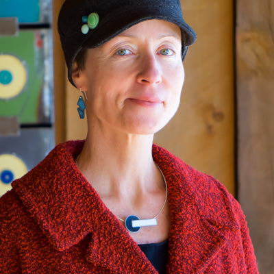 Wearing the Lana - Ford & Dodge necklace, a woman in a textured red coat, black hat with green pins, and blue geometric earrings stands indoors, smiling softly with vibrant artwork blurred behind her. Handmade jewelry by Joyride Jewelry.
