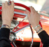 Close-up of a person’s hands on a vintage red steering wheel in an Emril - Ford & Chevy, wearing multiple silver rings and colorful stone bracelets. Dashboard and steering column are visible in the background.