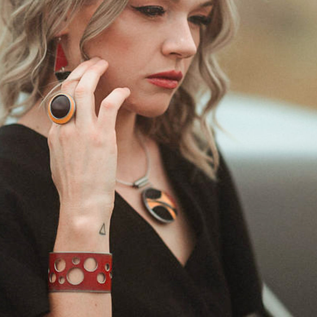 A woman with wavy blonde hair, red lipstick, and a black outfit poses . She wears geometric jewelry: an adjustable ring, red cuff bracelet with cutouts, triangular earrings, and a statement necklace all by Joyride Jewelry