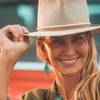 A smiling woman outdoors tilts her beige wide-brimmed June - Ford hat with one hand. She wears turquoise earrings with sterling silver ear wires and has long blonde hair, with blurred red and blue hues in the background.