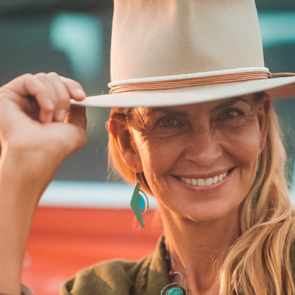 A smiling woman outdoors tilts her beige wide-brimmed June - Ford hat with one hand. She wears turquoise earrings with sterling silver ear wires and has long blonde hair, with blurred red and blue hues in the background.