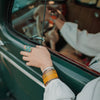 Wearing the Frankie - Dodge pickup adjustable bracelet by Joyride Jewelry, a person rests their hand on the open window of a vintage green Ford car, with another hand on the steering wheel showcasing the classic car’s original paint.